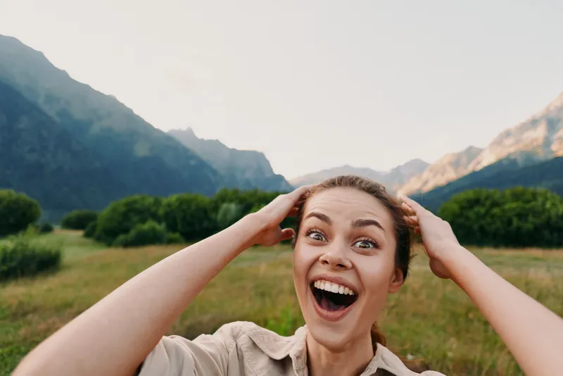 Happy woman with hands on head smiling outdoors in nature with mountains and green bushes in the background. Concept of lifestyle, joy, and freedom in natural environment.