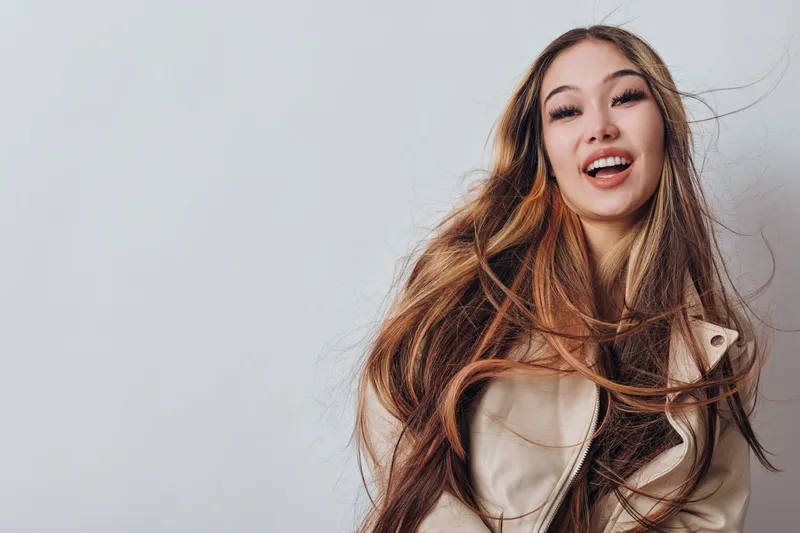 Young woman with long hair smiling and wearing light jacket on white background. Portrait of happy female with natural makeup and casual hairstyle indoors with positive attitude.