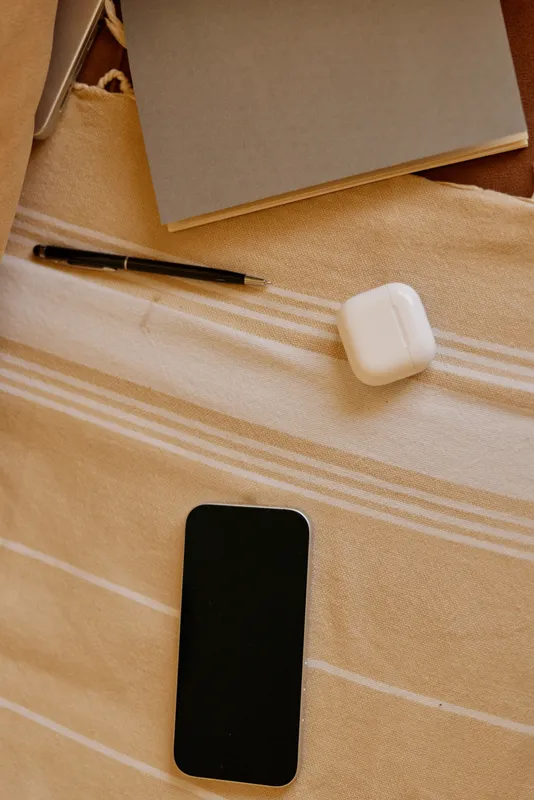 A cozy bedroom scene with a smartphone, notebook, pen, and earbuds on a striped blanket beside a soft pillow, capturing a home productivity moment and calm planning.
