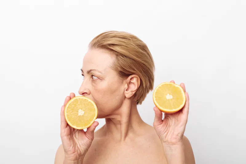 Mature woman with short hair holds two halves of a fresh orange near her face on white background. Concept of natural skincare, healthy lifestyle, vitamin C, and wellness for aging skin.