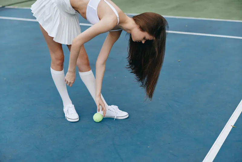 Young female tennis player in a white tennis outfit, picking up a green ball on a blue court, exuding concentration and athleticism during a match