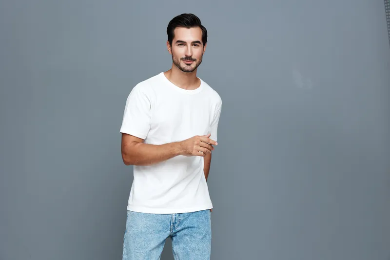 Confident young caucasian man wearing a white t-shirt, standing and smiling at the camera while holding a hand gesture, isolated on a gray background studio portrait.
