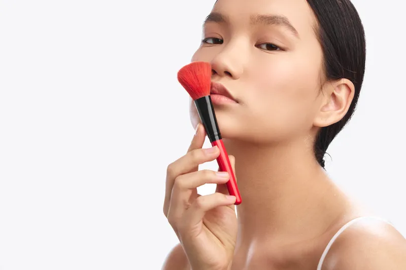 Asian woman applies makeup with a brush in a bright studio, focusing on skincare and beauty routine. The scene conveys elegance, confidence, and a modern cosmetic ritual.