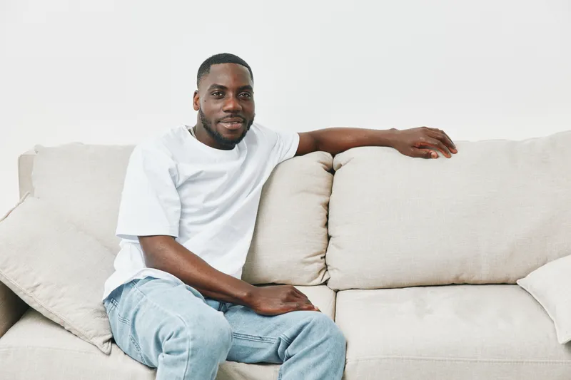Relaxed African American man in casual white t shirt sitting on a light beige couch, exuding a friendly and approachable vibe, perfect for lifestyle themes