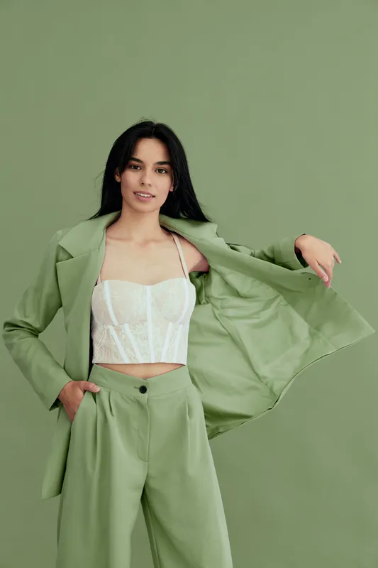 young woman wearing green pantsuit and white lace corset jacket poses confidently with flowing coat against plain green background, stylish and modern look