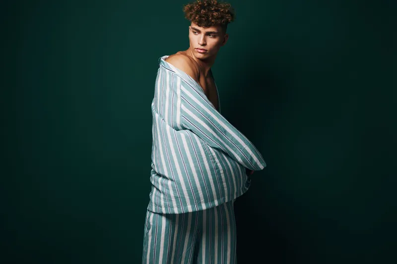 Young man wearing striped pajamas standing against dark background looking sideways with thoughtful expression and casual posture in studio lighting.