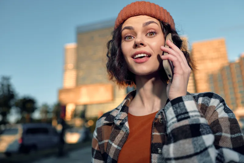 Woman on phone in urban street, candid golden hour glow portrait with beanie and smile, outdoor lifestyle conveying authenticity, mindful living and emotional storytelling.