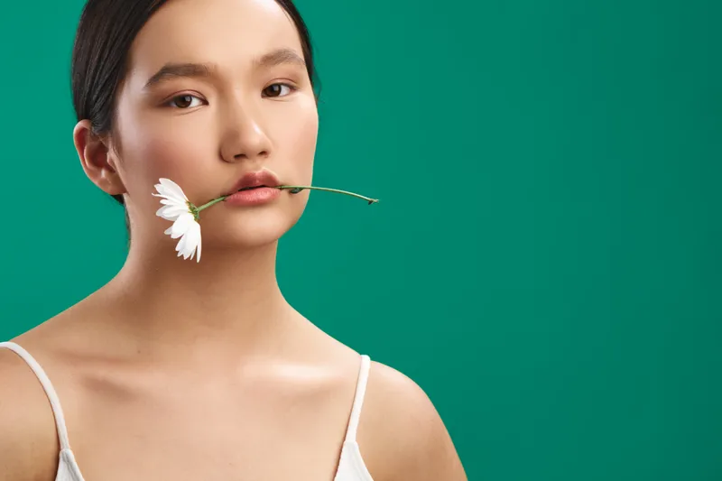asian woman softly poses with a white flower held in her lips, minimal portrait against a bold green backdrop, conveying calm beauty and delicate poise, artistic mood, clean composition, contemporary