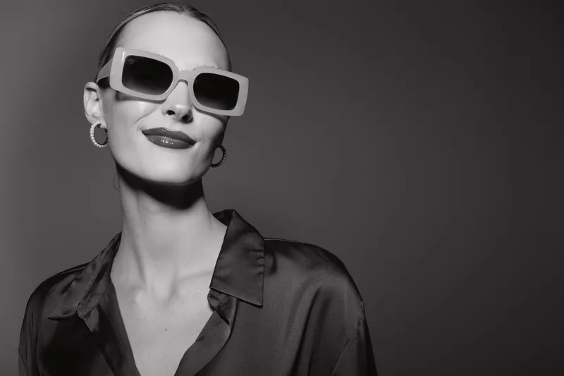Elegant white woman with pale skin wearing large sunglasses and hoop earrings poses confidently in a black satin shirt against a dark background. Concept of fashion and style.