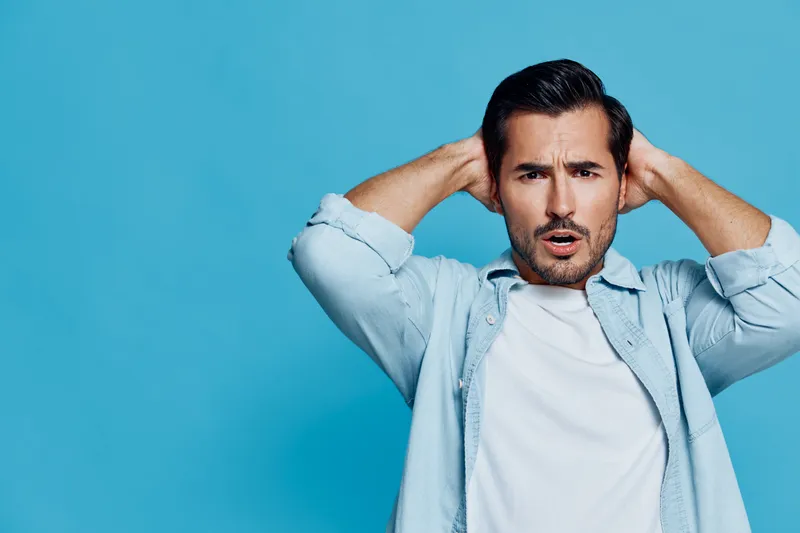 Surprised man with short hair wearing a light blue shirt holds his hands behind his head, gazing at the camera against a solid blue background. Expression of shock and disbelief.