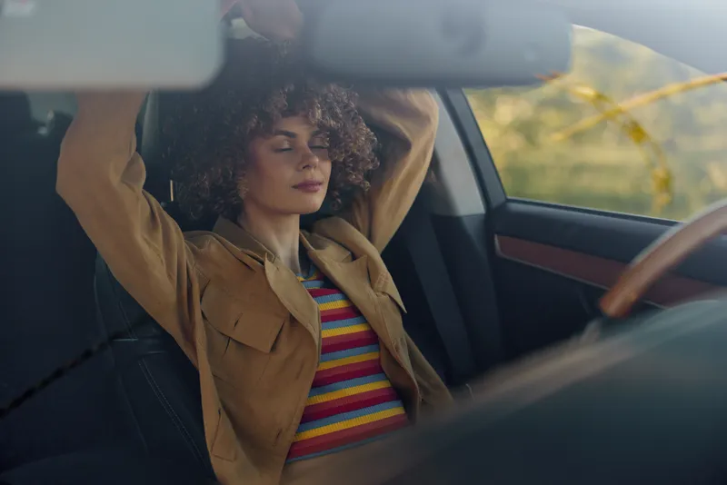 Relaxed woman with curly hair sits in a car interior wearing a striped shirt and tan jacket, eyes softly closed, enjoying a peaceful moment. Retro mood and travel vibe evoke self care.