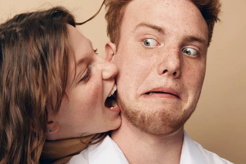 A playful young woman pretends to bite a surprised young man with freckles in a close-up studio portrait on beige background. Concept of fun, friendship, and humorous interaction.