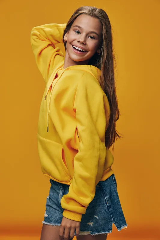 Teen girl smiling and posing in yellow hoodie and denim shorts on orange background with casual style and long hair in joyful mood.