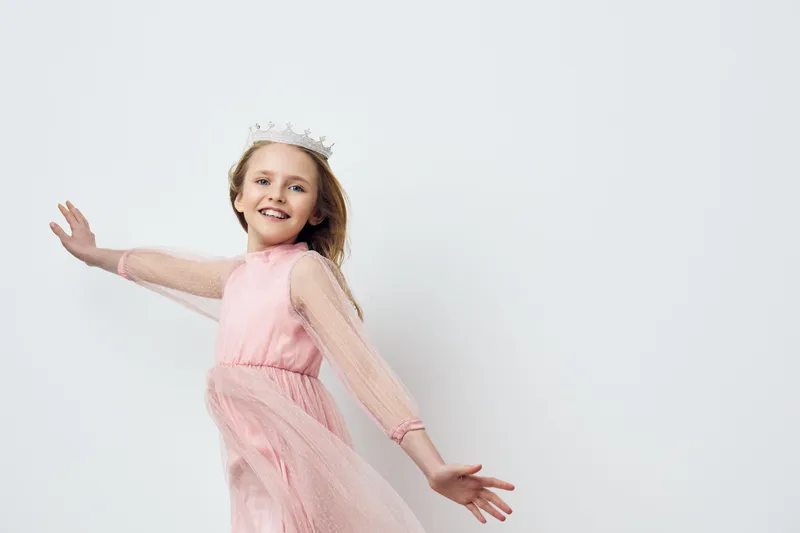 Joyful young girl wearing a pink dress and a crown, dancing with a smile against a minimalistic white background, perfect for concepts of childhood happiness and celebration