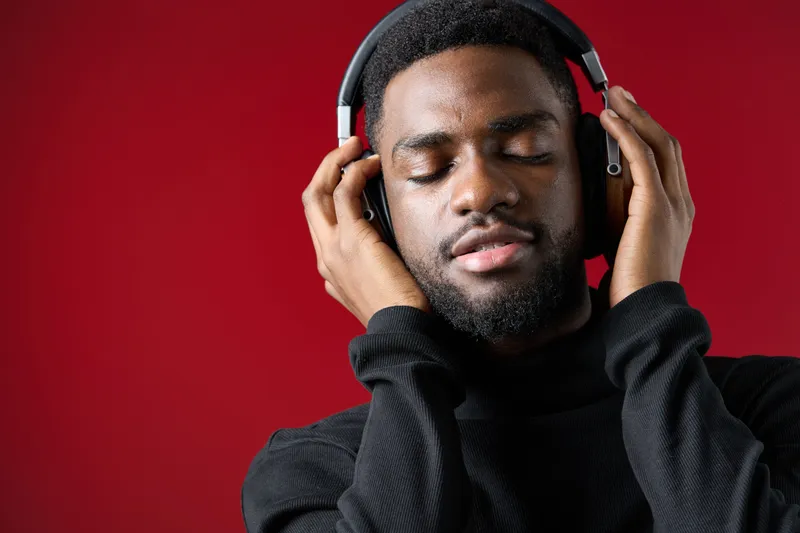 Young Black man enjoying music with headphones, immersed in sound against a red background, expressing joy and relaxation in a stylish black sweater