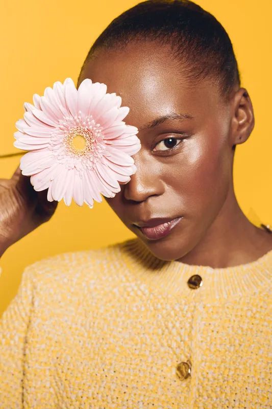 Close-up of a dark skinned African American woman holding a pink flower covering one eye. She wears a yellow knitted sweater against a bright yellow background. Concept of beauty and nature.