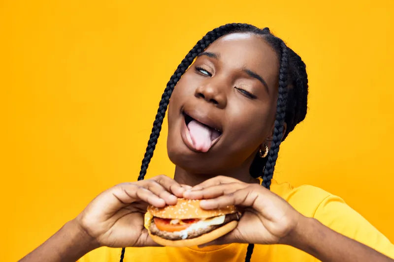 Young woman playfully enjoying a burger against a vibrant yellow background, showcasing fun emotions and a casual style with braids and a bright t shirt