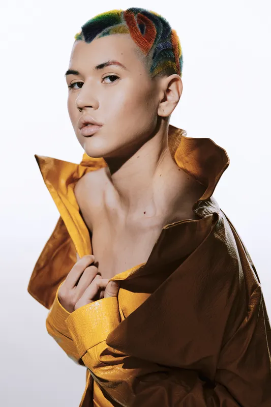 Young woman with colorful braided hair wearing a mustard yellow jacket, striking confident pose against white background, modern and stylish portrait lighting.