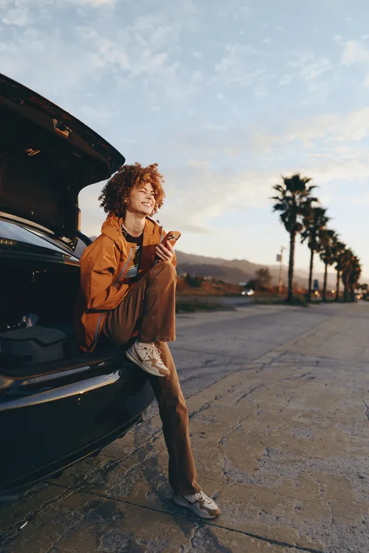 Woman lifestyle near an open car trunk on a quiet road at sunset, holding phone and jacket. Outdoor travel moment by palm trees, cinematic backlight, casual mood today.