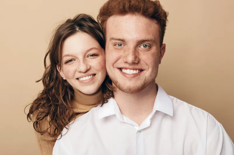 Smiling young couple with curly hair and red hair posing together on beige background. Happy man and woman show positive emotions and close connection in casual clothing. Concept of love and