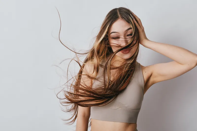 Young woman with long flowing hair wearing beige tank top posing with eyes closed and hand near face against plain light background in studio for beauty and fashion portrait.