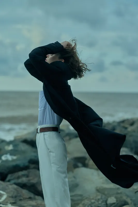 Wind, coat, fashion, seaside, movement, windy scene of a model by the ocean on a rocky shore with flowing garment and breeze