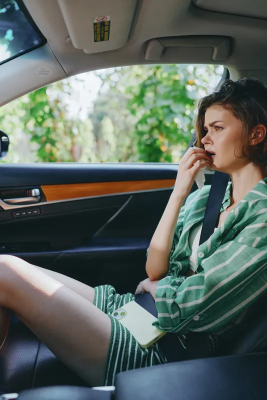 Young woman sits in a car seat with a worried expression, biting her finger while wearing a green striped shirt, natural light, casual travel moment.
