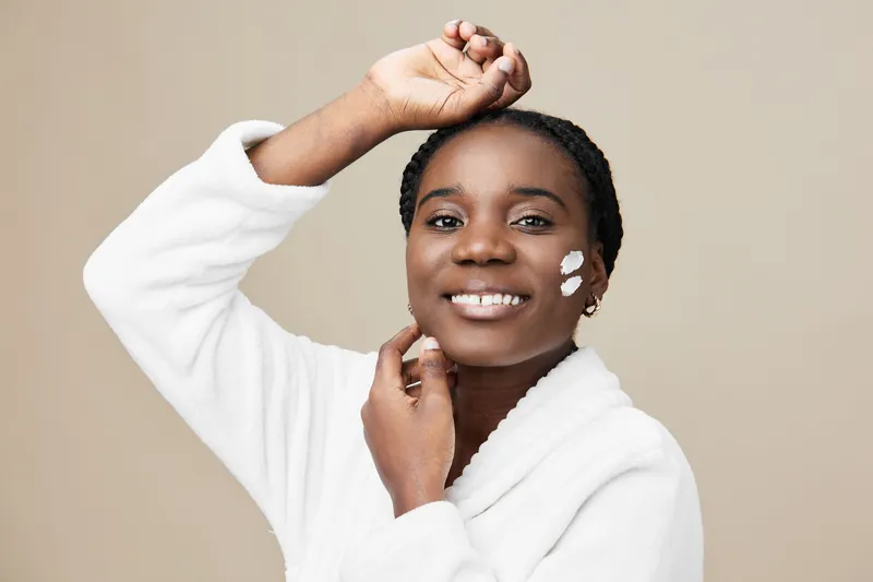 Happy Black woman applying facial cream, smiling brightly in a cozy white robe against a soft beige background, emphasizing skincare and self care routines