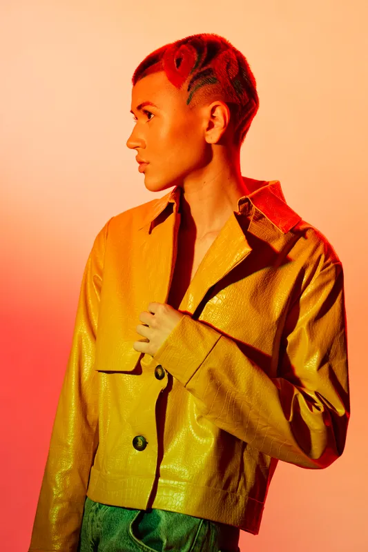 Young woman with creative red shaved hairstyle wearing a shiny yellow jacket poses against warm orange background, showcasing bold fashion and modern style.