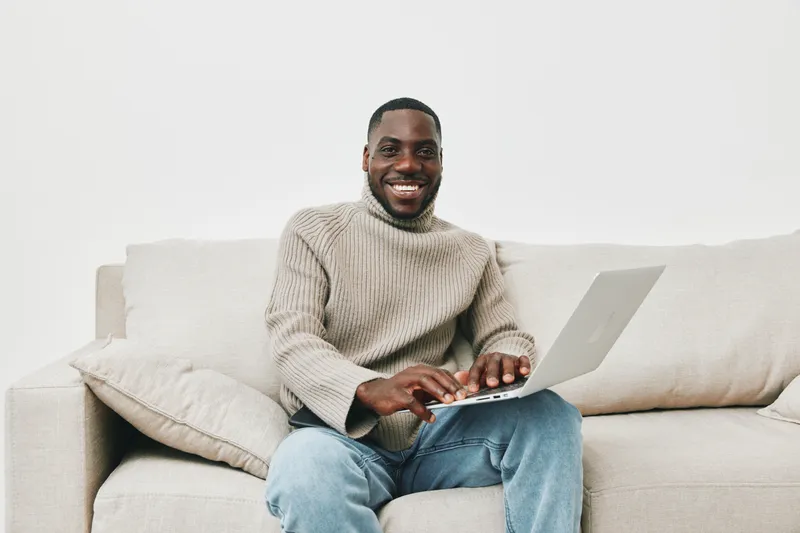 Cheerful young Black man working on a laptop at home in casual knitwear, embodying comfort and productivity in a modern living space