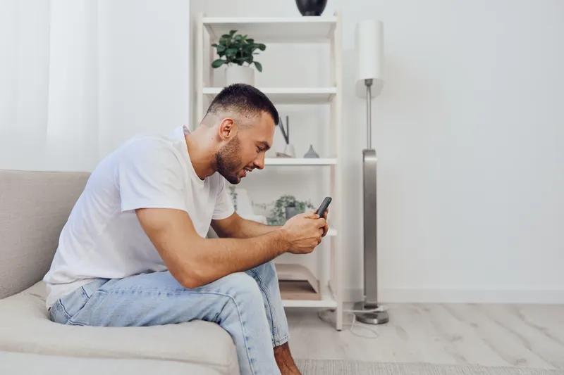 Thoughtful man using smartphone on a cozy couch, reflecting modern lifestyle and technology trends. A serene indoor setting with soft colors enhances the mood.