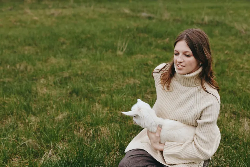 Goat, kid, woman, outdoors, field, grass, lifestyle, animal, pet, nature a woman in a cream sweater gently holds a white goat kid in a green field, a calm wholesome lifestyle moment captured outdoors