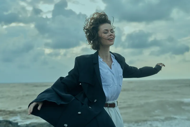 Wind, beach, coat, business attire set the scene for a confident, free moment by the ocean, with windblown movement and outdoor breeze. A lively, relaxed look conveys empowerment, style, and ambition