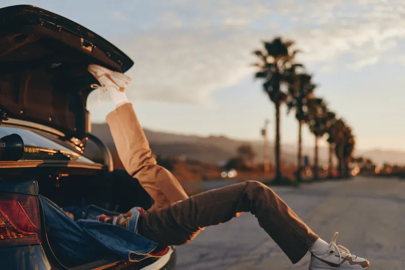Man lifestyle scene with car trunk open during sunset, road trip vacation mood, seated legs stretching outward, casual jacket and sneakers, palm highway scenery, calm reflective travel