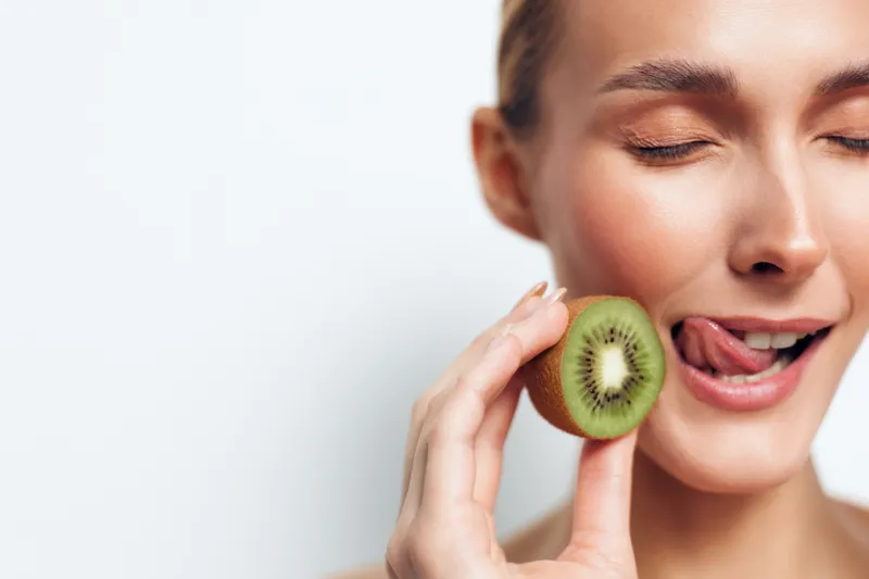 Woman holding kiwi fruit slice close up smiling healthy skincare concept, fresh face on light background, fingers touch the fruit to her lips, natural beauty wellness