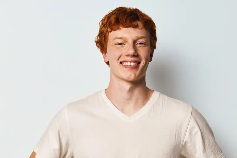 Young man with red hair and fair skin smiling looking at camera in white t shirt with plain background casual confident expression.