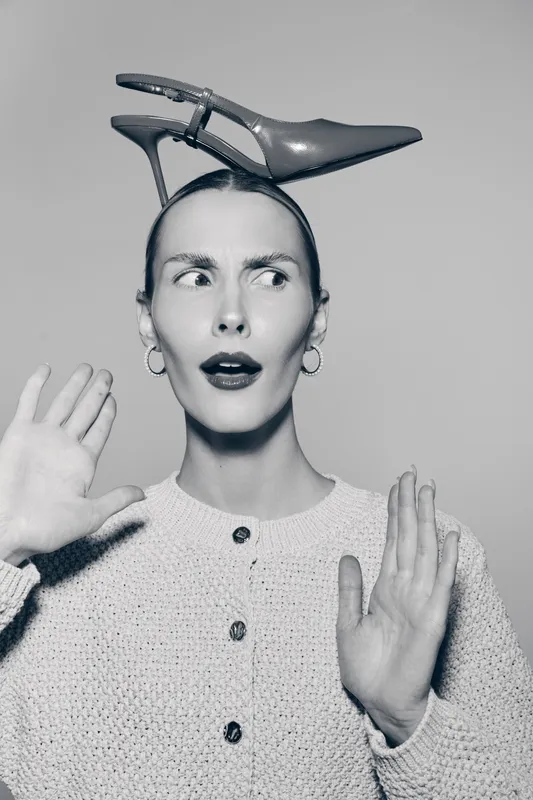 Young white woman with light skin and red lips wears beige sweater and holds hands up while balancing high heel shoe on head. Concept of fashion, fun, and quirky style.