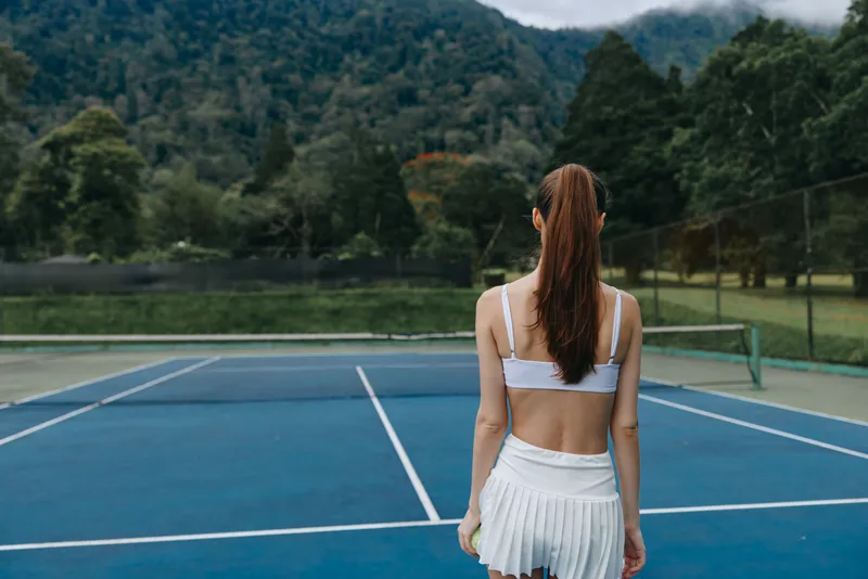 Young female tennis player in a white outfit standing on a tennis court, showcasing determination The backdrop features lush green mountains and a blue court