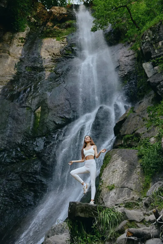 young woman practicing yoga in white sportwear stands on rock near tall waterfall surrounded by green forest and mossy stones outdoor nature calm energy