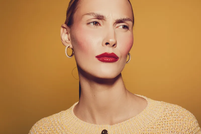 Close-up portrait of a woman with red lipstick and hoop earrings wearing a beige sweater on a warm yellow background. Concept of fashion, beauty, and confident expression.