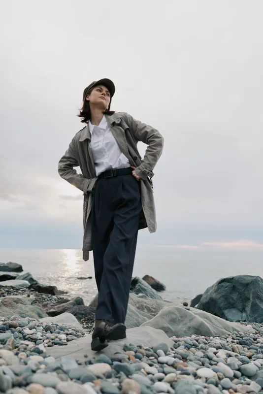 Young woman in casual outfit stands confidently on rocky beach near calm sea under cloudy sky. Concept of fashion, freedom, nature, and outdoor lifestyle in cool weather conditions.