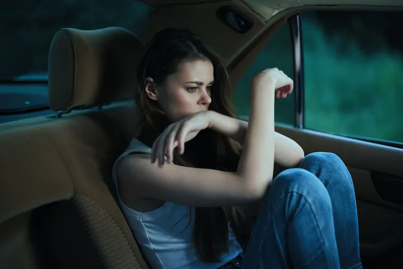 Pensive young woman sitting alone in a car, reflecting on life, wearing a casual white tank top and blue jeans, with a moody nighttime background.