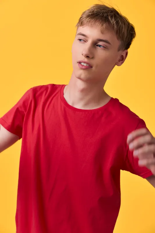 Man in a vibrant red shirt poses casually against a bright yellow studio backdrop, captured in a dynamic moment with relaxed stance and soft lighting that highlights youthful style and modern fashion.