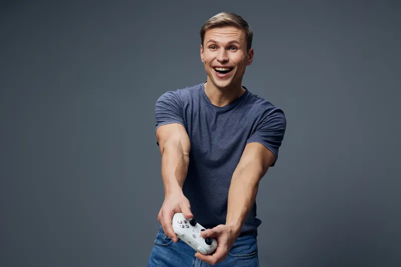 Full body joyful young caucasian man in casual blue t-shirt playing video game with white controller, smiling broadly, excited expression, against plain gray background. People entertainment lifestyle