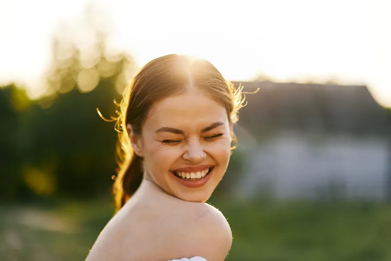 Smiling young woman with long hair enjoying a sunny day, wearing a white dress and showing joy against a bright outdoor background with lush greenery