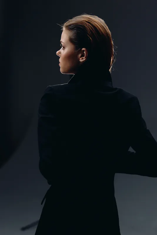 Profile of a thoughtful woman in a black coat against dark background, with dramatic lighting and elegant style.