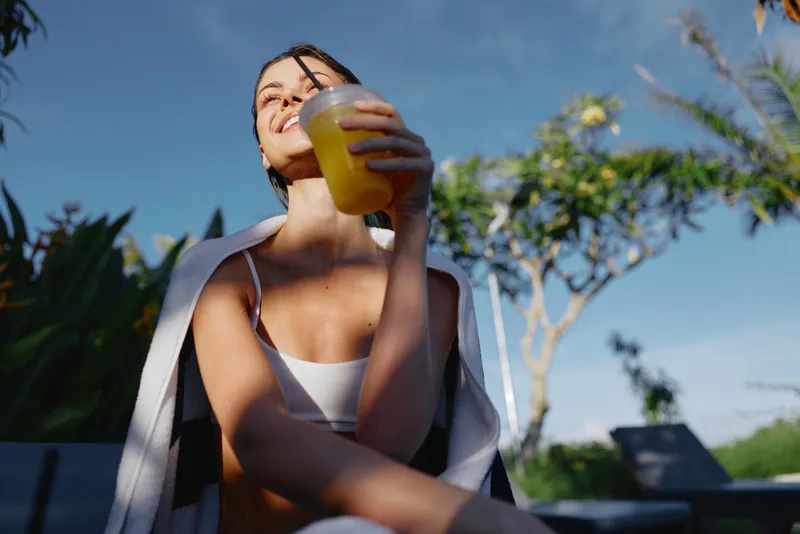 A relaxed outdoor moment with a smiling woman enjoying a refreshing beverage under bright sunshine. A lifestyle scene focused on hydration, wellness, and everyday joy.