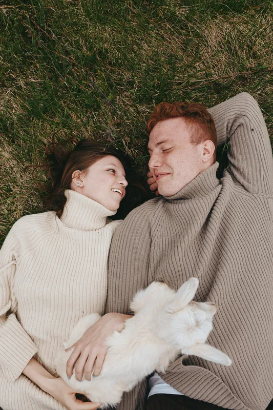 Lifestyle couple lying on grass in cozy sweaters with a goat kid nearby, sharing warm smiles in an outdoor nature setting, a tender moment of affection and calm companionship.