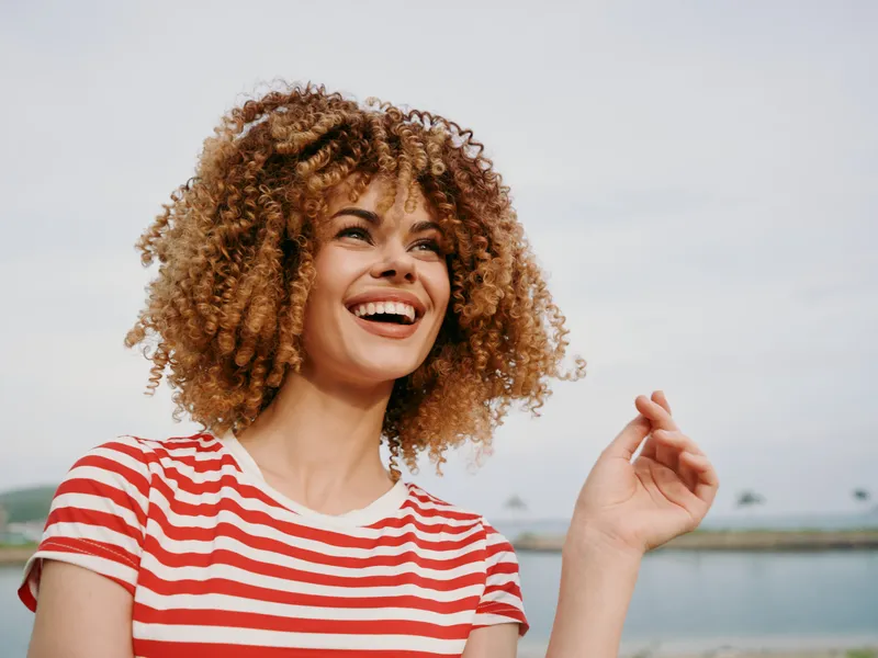 A happy young woman with curly hair in a red and white striped shirt stands outdoors by a beach, smiling brightly as sunlight highlights her joyful expression.