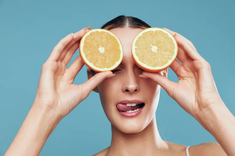 Young woman holding two orange halves in front of her eyes, playful expression, tongue out, smooth skin, bright background, fresh fruit, healthy lifestyle concept